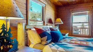Un dormitorio de cabaña de madera con una cama, almohadas, una mesita de noche, una lámpara y una ventana.