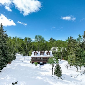 A cabin with a snow-covered roof, surrounded by trees and a clear sky.
