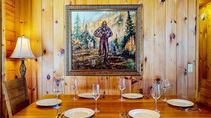 Una mesa de comedor dispuesta con platos y copas de vino, un cuadro enmarcado de un paisaje forestal, y paredes de madera.