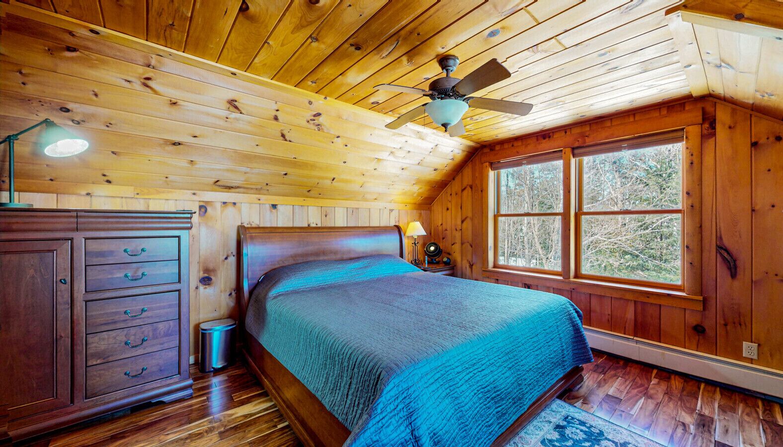 Ein Schlafzimmer einer Holzhütte mit einem Bett, einem Schminktisch und einem Fenster mit Blick auf Bäume.
