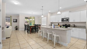 A modern kitchen with white cabinets, a central island, and a dining area with chairs. - Cuban Palms Paradise | Theatre & Games - Luxe Resort + 10 Min Disney + Pet Friendly (Kissimmee)
