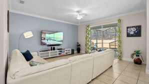 A living room with a large flat-screen TV, a ceiling fan, a sofa, a TV stand, and a sliding glass door leading to an outdoor area. - Cuban Palms Paradise | Theatre & Games - Luxe Resort + 10 Min Disney + Pet Friendly (Kissimmee)