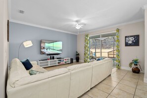 Ein Wohnzimmer mit großem Flachbildfernseher, Deckenventilator, Sofa, TV-Schrank und einer Glastür, die zu einem Außenbereich führt.