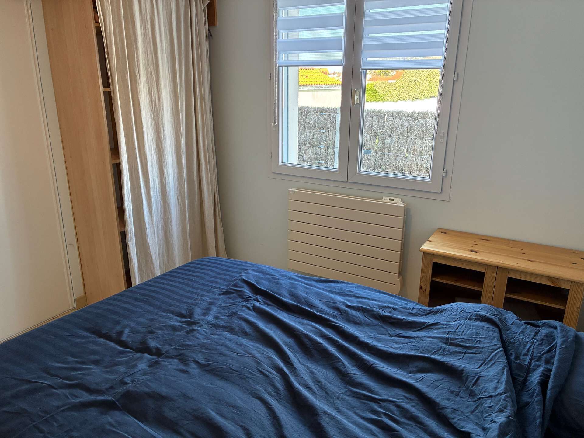 A bedroom with a bed, a window with blinds, a radiator, and a wooden bench.