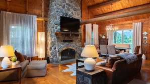 A log cabin interior with a stone fireplace, wooden furniture, and a television mounted on the wall. - Scenic, Dog-Friendly Log Cabin: Minutes to Lake Winnipesaukee, Gunstock Mountain (Gilford)