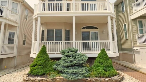 Une maison à deux étages dotée d’un balcon, d’un porche et d’un petit jardin où poussent trois arbres à feuilles persistantes.