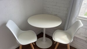 Ett litet, runt, vitt bord med två vita stolar med ben av trä, placerat framför en vit tegelvägg med ett fönster.