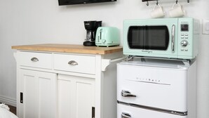 Ett litet kök med en mikrovågsugn, brödrost och kaffebryggare placerade på ett vitt skåp, en TV som är monterad på väggen och en hylla med koppar.