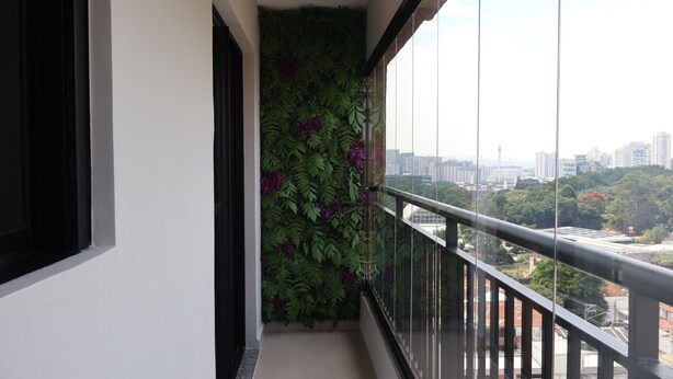 Un balcón con baranda de vidrio, vistas a la ciudad y una pared verde con flores moradas.
