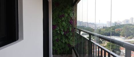 A balcony with a glass railing, a cityscape view, and a green wall with purple flowers.