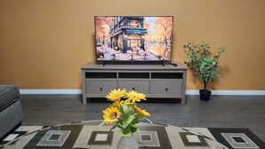 Ein Wohnzimmer mit einem Flachbildfernseher auf einem Schrank, einer Couch, einem Couchtisch mit einer Vase voller Sonnenblumen und einer Topfpflanze.
