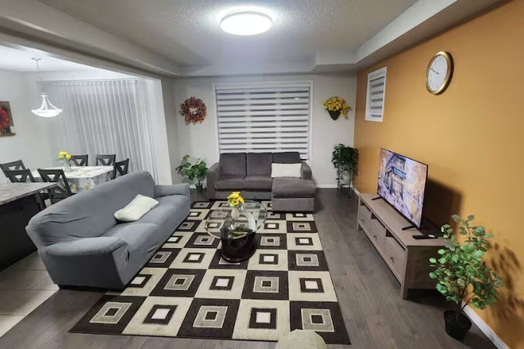 Ein Wohnzimmer mit grauem Sofa, einem gemusterten Teppich, einem Couchtisch, einem Fernseher und einem Essbereich mit Tisch und Stühlen.