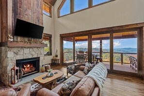Un salón amplio con chimenea de piedra, un televisor de pantalla plana grande y vistas a un paisaje montañoso a través de amplios ventanales.