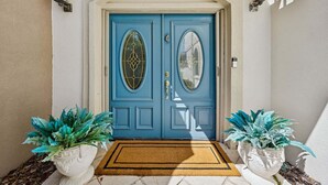 A blue double door with glass panels, flanked by two potted plants, and a doormat on the ground. - Private pool, 3-bedrooms, 3.5 bathrooms, 2-car garage with EV charger, across from beach access (Fernandina Beach)