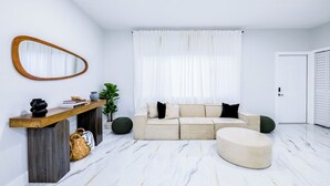 A modern living room with a sectional sofa, a round ottoman, a wooden console table, a mirror, and a potted plant. - HARD ROCK OASIS - 3BR Pool Home + Game Room (Miami)