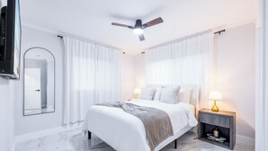A bedroom with a bed, a nightstand, a ceiling fan, a mirror, and a door. - HARD ROCK OASIS - 3BR Pool Home + Game Room (Miami)