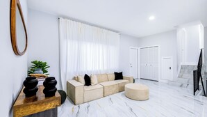 A modern living room with a sectional sofa, a round ottoman, a wooden coffee table, and a large mirror. - HARD ROCK OASIS - 3BR Pool Home + Game Room (Miami)
