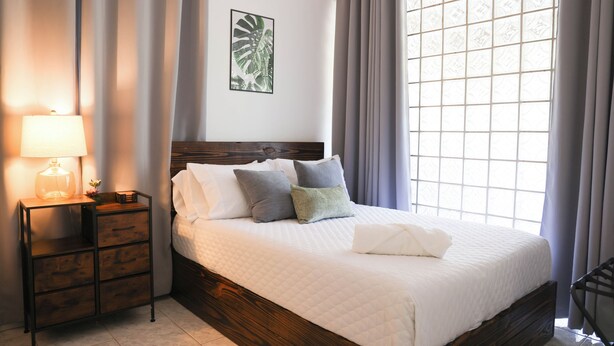 A neatly made bed with pillows, a bedside table with a lamp, and a window with curtains. - Hacienda Yagrumo - Spacious 4-bedroom house - Mountain Retreat with River (Utuado)