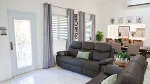 A living room with a sofa, a dining area with chairs, a kitchen in the background, and a large window with curtains. - Hacienda Yagrumo - Spacious 4-bedroom house - Mountain Retreat with River (Utuado)
