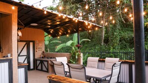 An outdoor patio with a table and chairs, string lights, and a view of greenery. - Hacienda Yagrumo - Spacious 4-bedroom house - Mountain Retreat with River (Utuado)