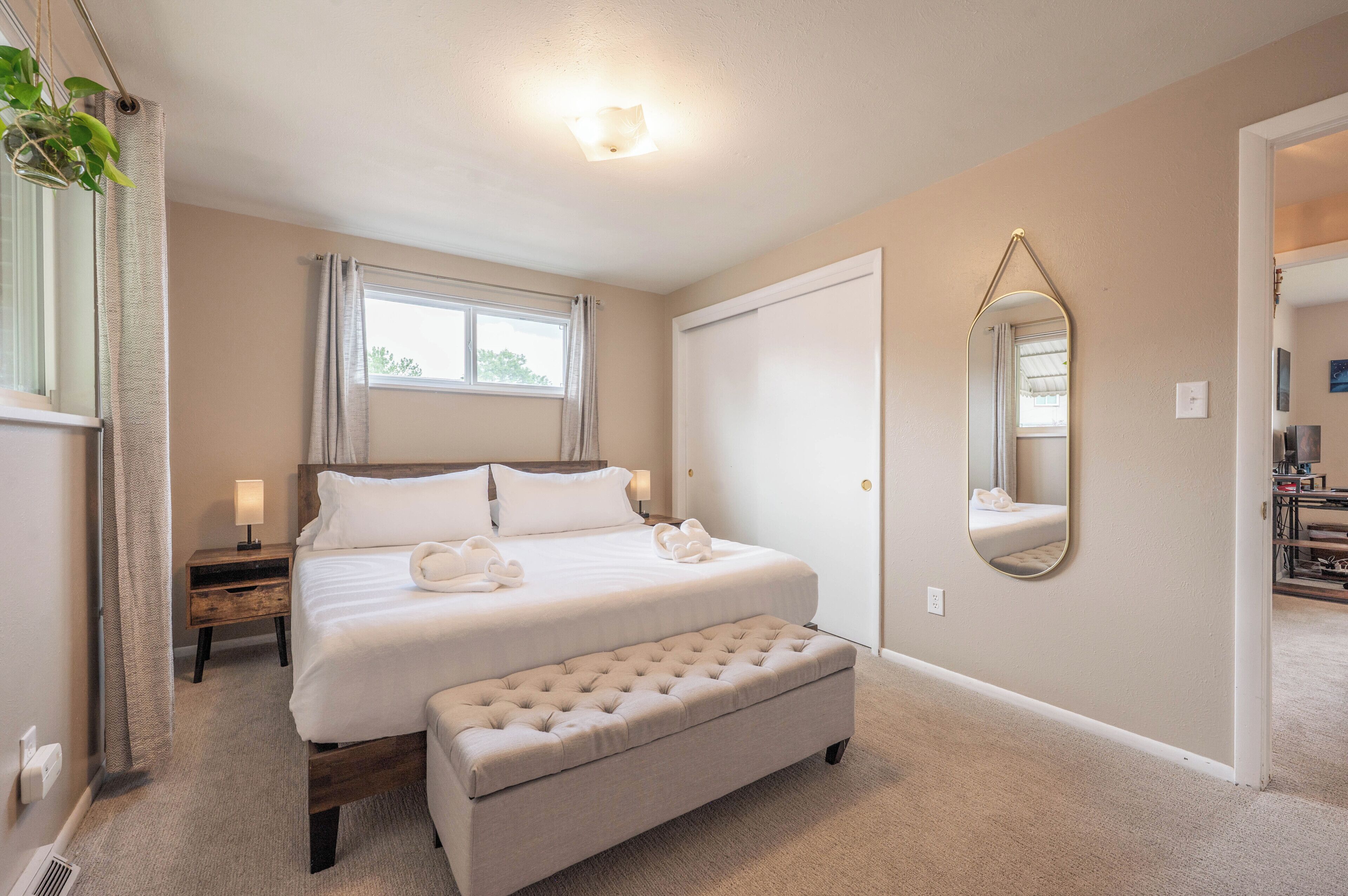 A hotel room with a bed, a bench, a bedside table with a lamp, a window with curtains, a door, and a mirror on the wall.