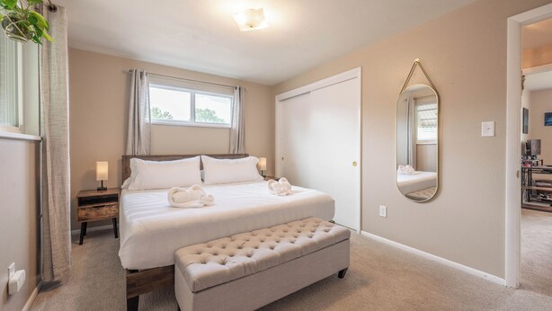 A hotel room with a bed, a bench, a bedside table with a lamp, a window with curtains, a door, and a mirror on the wall. - Golden Home in the Valley near Red Rocks (Golden)