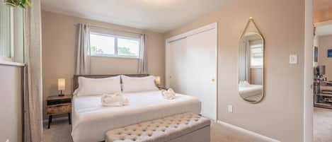Ein Hotelzimmer mit Bett, Sitzbank, Nachttisch mit Lampe, Vorhängen am Fenster, einer Tür und einem Spiegel an der Wand.