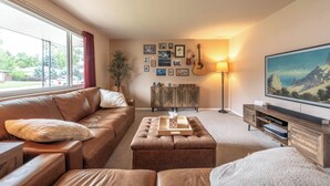 A living room with a brown leather sofa, a wooden coffee table, a TV stand, and a large window with a view of trees. - Golden Home in the Valley near Red Rocks (Golden)