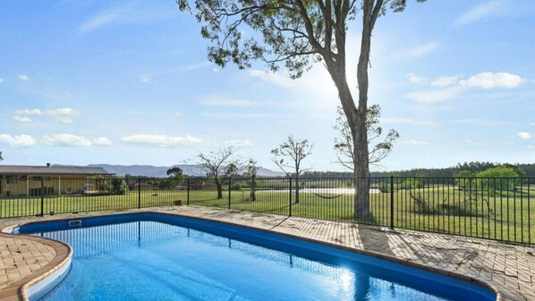 A swimming pool with a paved deck, surrounded by a black fence, and a large tree in the background.
