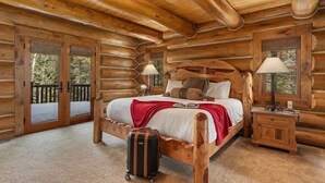 A log cabin bedroom with a wooden bed, two bedside lamps, a suitcase, and a balcony with glass doors. - Abode at Forest Edge, luxury log home, hot tub (Wilson)