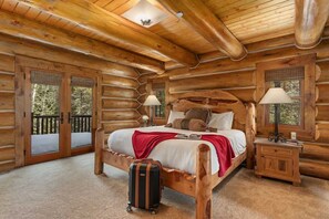 A log cabin bedroom with a wooden bed, two bedside lamps, a suitcase, and a balcony with glass doors. - Abode at Forest Edge, luxury log home, hot tub (Wilson)