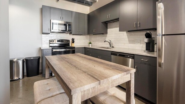 A modern kitchen with stainless steel appliances, dark cabinetry, and a wooden table. - Landing | Spectacular 2BD in Uptown (Memphis)