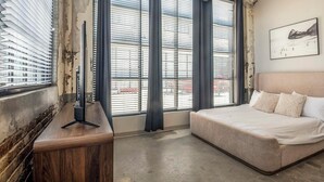 A bedroom with a bed, a desk with a monitor, large windows with blinds, and a painting on the wall. - Landing | Spectacular 2BD in Uptown (Memphis)