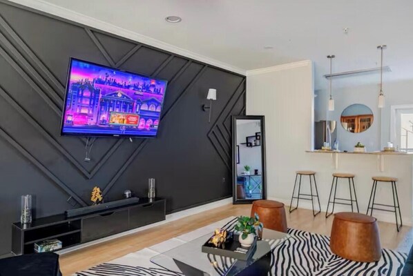 A modern living room with a flat-screen TV mounted on a dark wall, a coffee table with decorative items, and a bar area with stools.