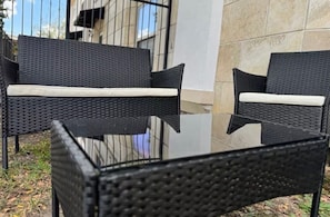 Two black wicker chairs with white cushions on a glass table, set against a building with a tiled wall.