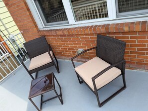 Two wicker chairs with cushions on a balcony with a brick wall and a window in the background.