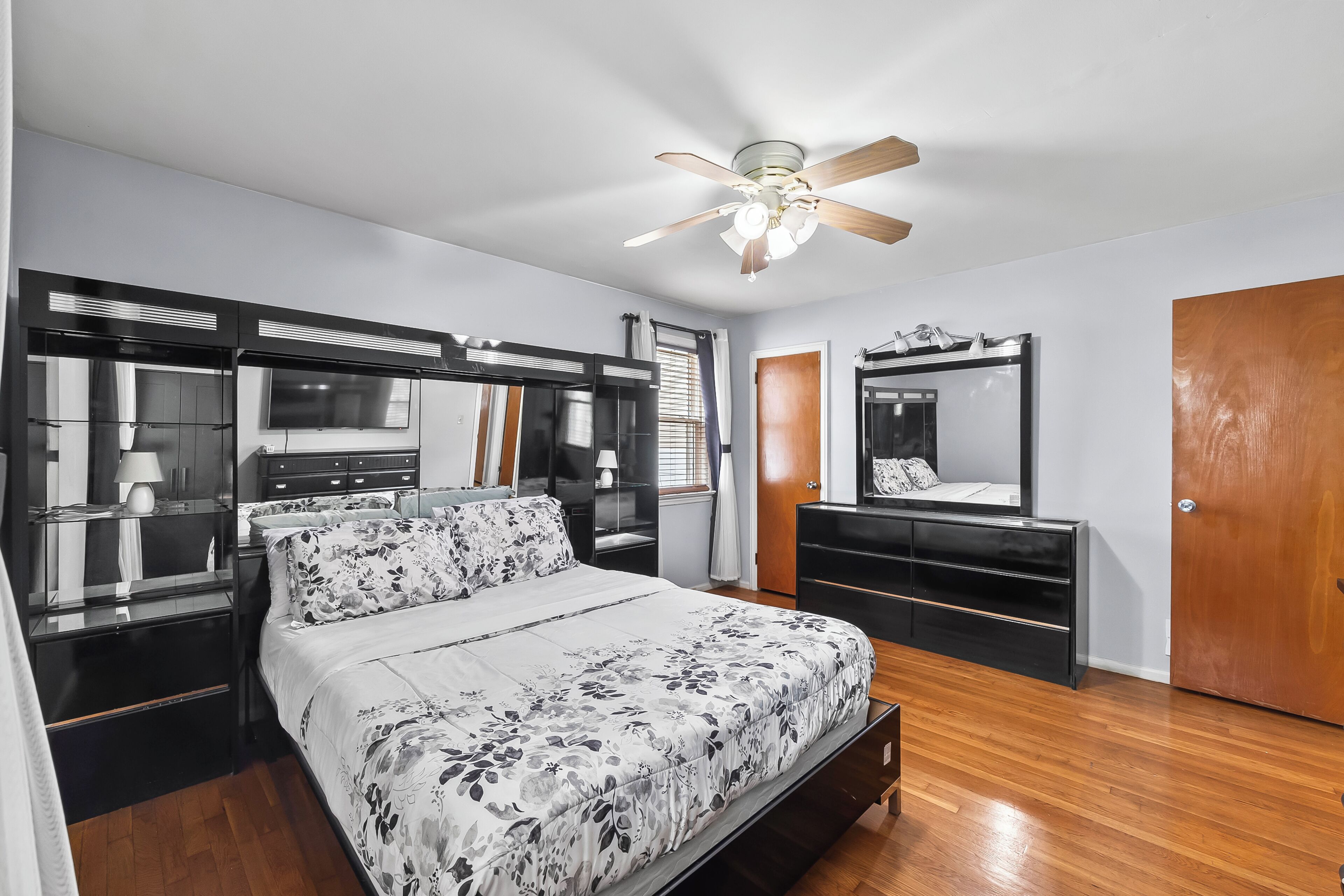 Un dormitorio con cama, armario, cómoda, espejo, ventilador de techo y una puerta.
