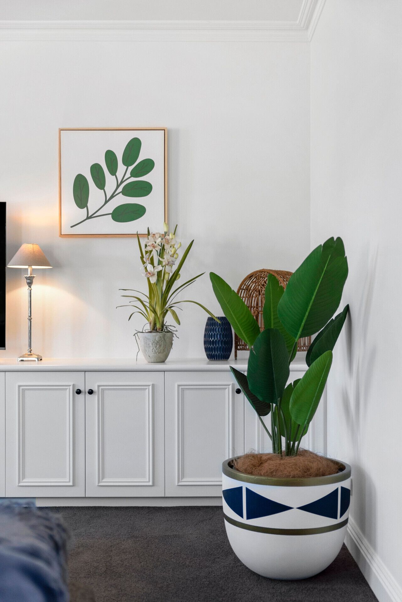 Ein Wohnzimmer mit einem weißen Schrank, einer Topfpflanze, einem gerahmten Kunstwerk und einer Lampe.