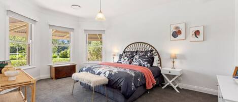 A bedroom with a large bed, two bedside tables, a chair, and a window with a view of greenery.