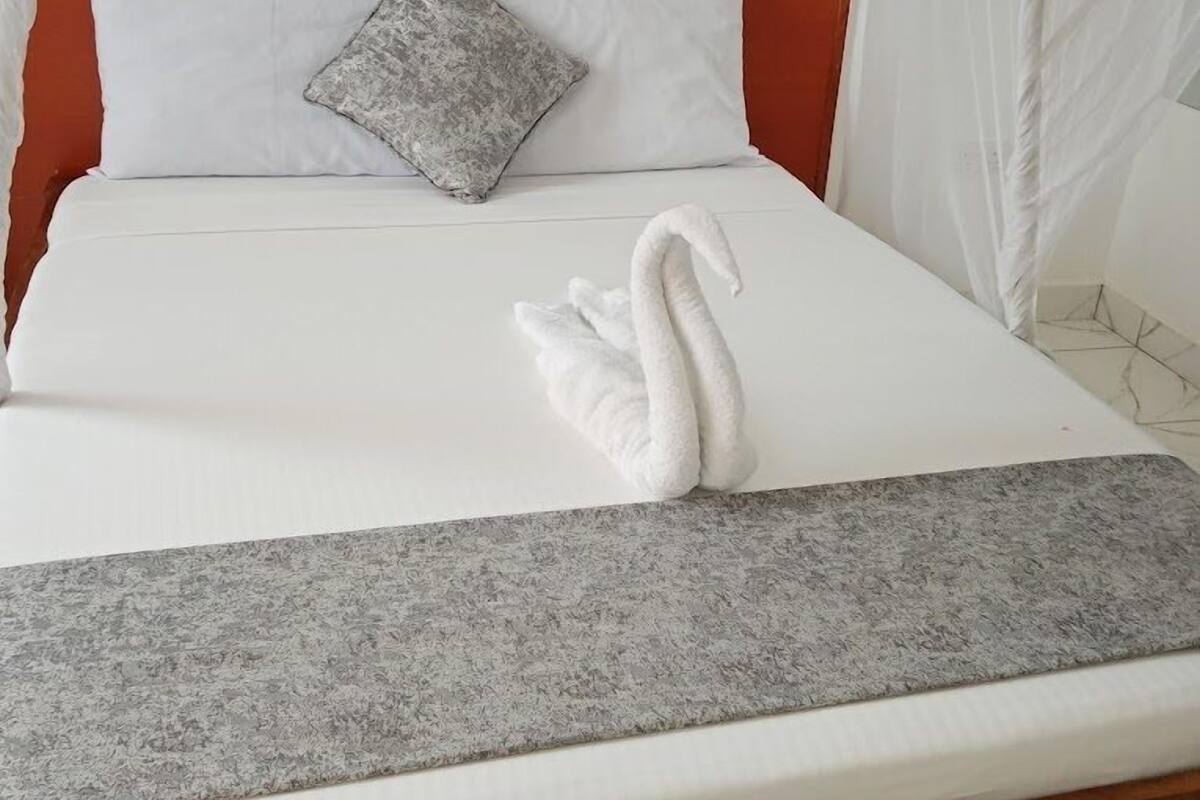 Una cama con un colchón blanco, un plaid gris y una toalla con forma de cisne.