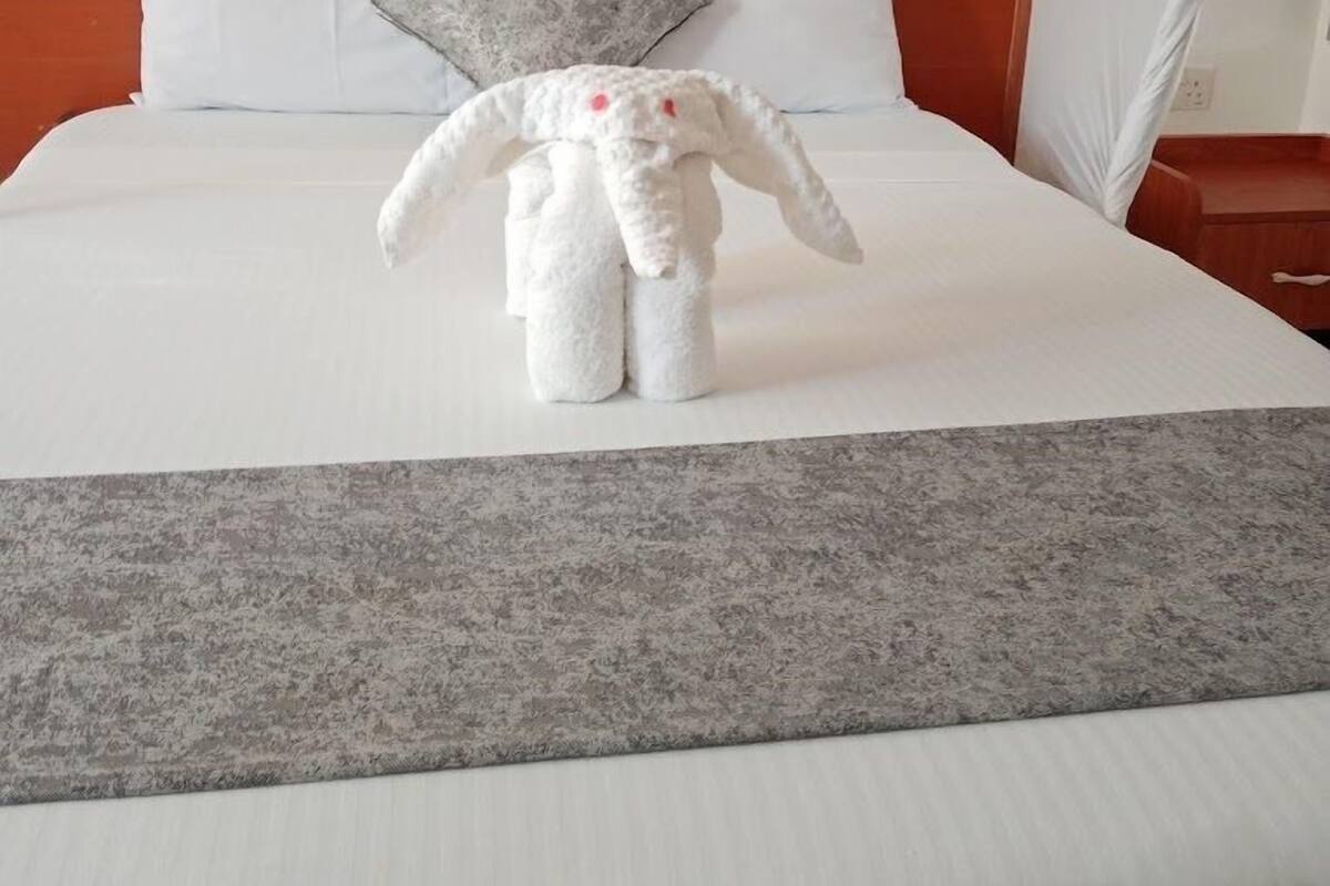 Habitación de hotel con cama, almohadas, una manta gris y una escultura de toalla blanca.