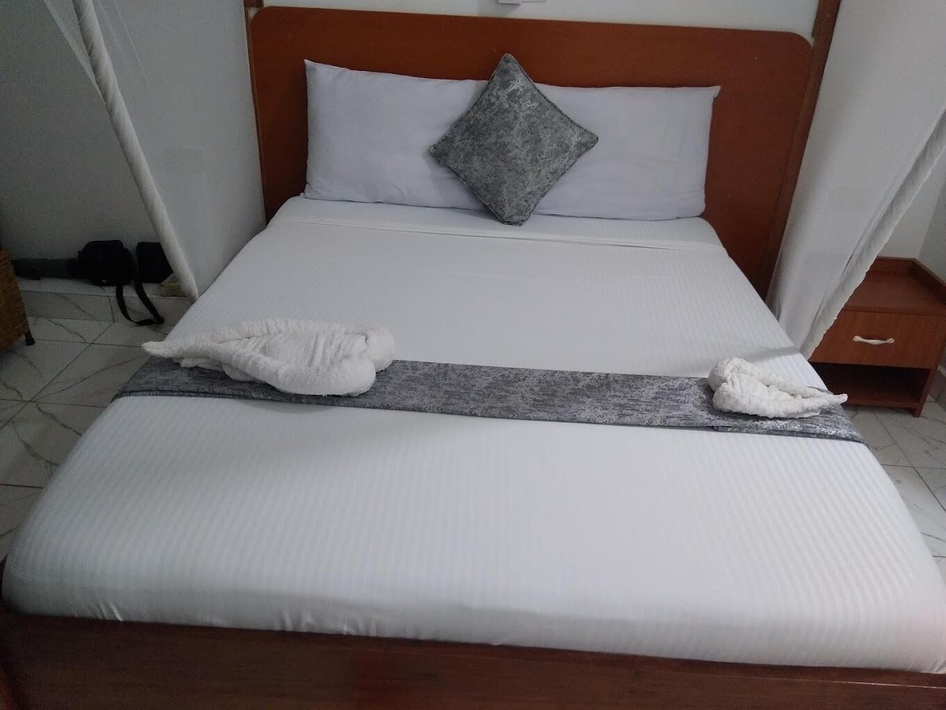 Una cama bien hecha con sábanas blancas, una almohada gris y dos toallas blancas sobre un cabecero de madera.
