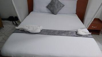 Una cama bien hecha con sábanas blancas, una almohada gris y dos toallas blancas sobre un cabecero de madera.
