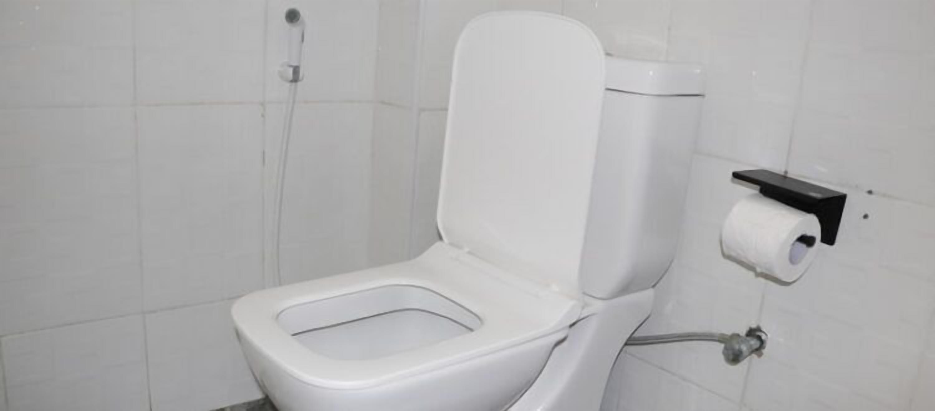 Inodoro blanco con tanque, dispensador de toallas de papel y un accionador de descarga en la pared.
