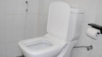 Inodoro blanco con tanque, dispensador de toallas de papel y un accionador de descarga en la pared.