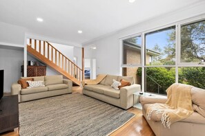 Un salón amplio con dos sofás, una escalera de madera y grandes ventanas que ofrecen vistas al verdor.
