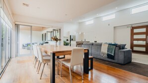 A spacious living area with a dining table, chairs, a sofa, and a large window. - Seabreeze Pacific Drive - Torquay - Sleeps 11! (Torquay)