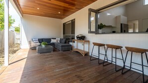 An outdoor patio with a wooden deck, a sofa, a coffee table, and bar stools. - Seabreeze Pacific Drive - Torquay - Sleeps 11! (Torquay)