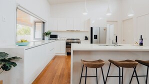 A modern kitchen with white cabinetry, stainless steel appliances, and a central island with stools. - Seabreeze Pacific Drive - Torquay - Sleeps 11! (Torquay)