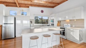 A modern kitchen with white cabinetry, a stainless steel refrigerator, a wooden ceiling, and a large window. - Ocean Views on Aldebaran - sleeps 11 (Ocean Grove)